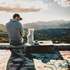 A man and his small white dog sitting together and looking out over the mountains